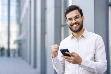 Gözlüklü genç adam elinde akıllı bir telefonla, şehir ortamında başarıyı kutluyor, mutluluk, güven ve modern teknoloji kullanımı yayıyor..