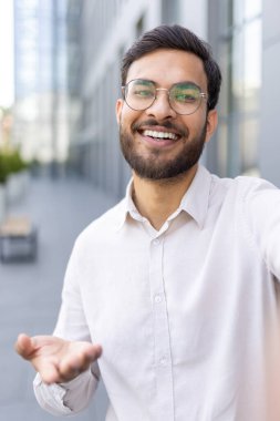 Modern bir şehirde, gözlüklü ve sakallı mutlu bir adam gülümsüyor. Onun anlamlı jestleri ve günlük kıyafetleri profesyonel ve olumlu temalar için ideal güven ve yaklaşımı iletir.