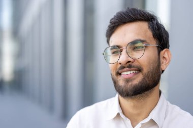 Genç bir adam, gözlük takarken güvenle gülümsüyor, şehir ortamında mutluluk ve rahatlık yayıyor..