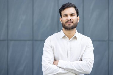Resmi giyinmiş genç bir adam, çağdaş mavi-gri arka plana karşı poz verirken kendine güvenini sergiliyor. Profesyonellik, güven ve başarı temaları için ideal.