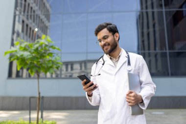 Laboratuvar önlüklü bir sağlık uzmanı dışarıda dururken akıllı telefonlarını kontrol ediyor. Sahne, tıbbi uygulamalarda modern teknoloji kullanımı ve profesyonel denge