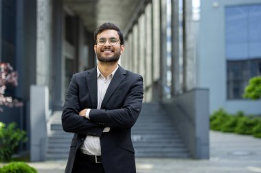 Kendine güvenen bir işadamı modern bir ofis binasının önünde duruyor, resmi bir takım elbise ve gözlük takıyor, elleri çapraz, olumlu ve profesyonel bir tavırla.