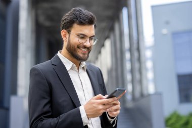 Resmi takım elbise giymiş gülümseyen bir adam şehir ortamında akıllı telefonunu karıştırıyor..