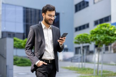 İş kıyafeti giymiş, nişanlı, akıllı telefonunu modern bir ofis binasının önünde kontrol ederken gülümseyen üretkenlik, bağlantı ve profesyonellik sergileyen kendine güvenen bir adam..