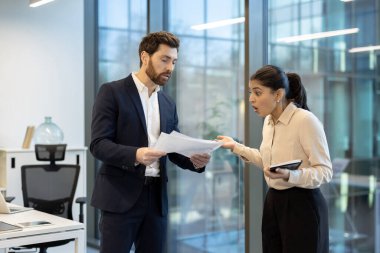 İki ofis çalışanı dokümanları incelerken ve modern bir çalışma alanında tablet kullanırken, işbirliği ve iletişimi göstererek canlı bir tartışmaya giriştiler.