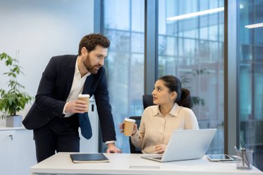 Çağdaş bir ofis ortamında etkileşime geçen iki iş arkadaşı, iş fikirlerini tartışırken aynı zamanda kahveyi paylaşıyor, takım çalışmasını ve iletişimi ön plana çıkarıyor.