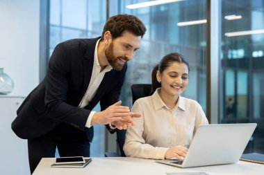 İki meslektaş, modern ofis ortamında bir dizüstü bilgisayarla iletişim kuruyor ve birlikte çalışıyor, takım çalışmasını teşvik ediyor, üretkenlik sağlıyor ve profesyonel ve kurumsal bir çevreye sahip olumlu bir çalışma ortamı sağlıyor.