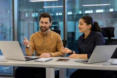 İki iş arkadaşı parlak, modern ofis ortamında dizüstü bilgisayarlar üzerinde çalışırken canlı bir tartışmaya giriştiler. Görüntü takım çalışmasını, iletişimi ve profesyonel bir atmosferde üretkenliği gösteriyor..