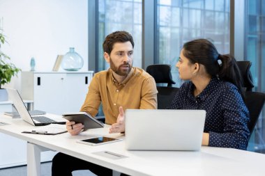 İki meslektaş, canlı bir çalışma alanında iş birliği yapıyor ve fikirleri paylaşıyor. Modern teknoloji ve dizüstü bilgisayarları birleştirerek etkili iletişim ve takım çalışmalarına odaklanırlar.