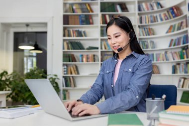 Profesyonel bir kadın kütüphanede oturur, kulaklık takar, dizüstü bilgisayarıyla çalışır, uzaktan destek sağlar ya da odaklanmış ve organize edilmiş bir ortamda çevrimiçi bir toplantı düzenler..