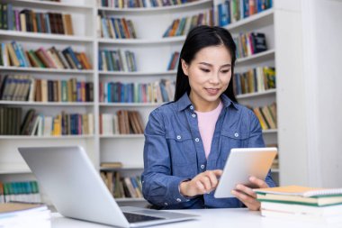 Genç bir kadın kütüphanedeki bir masada oturuyor, bir dizüstü bilgisayarın yanında dijital tablet kullanıyor. Çevre, düzenli ve odaklanmış bir ortamda öğrenmeyi veya profesyonel çalışmayı öneriyor.