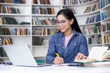 Genç bir kadın kütüphanede ders çalışıyor, dizüstü bilgisayarda çalışıyor ve kitaplarla çevrili notlar alıyor..