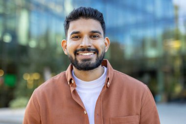 Hoş bir gülümsemesi olan genç bir adam modern cam bir binanın önünde pozitiflik ve güven atmosferi yaratıyor. Görüntü, kentsel bir hava ve doğal karizmayı yakalıyor..