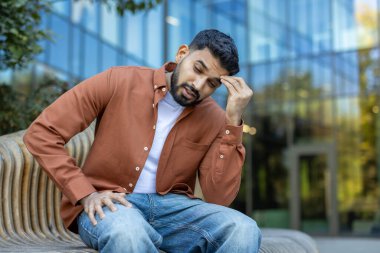 Sıradan giyinmiş bir adam, sıkıntılı bir ifadeyle kafasını tutup, modern bir binanın arka planına yaslanmış bir bankta oturuyor..