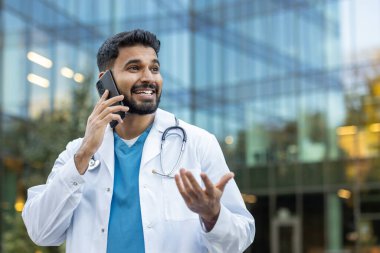Neşeli bir doktor dışarıda cam binanın arka planında telefonla konuşuyor..
