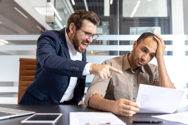 Hüsrana uğramış bir patron ofisteki bir belgeye işaret ederken bir çalışana bağırır.