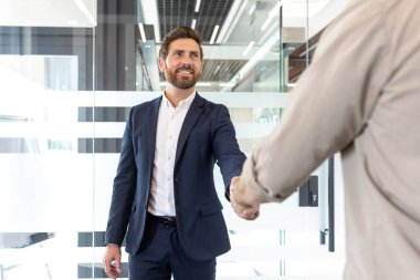 Modern bir çalışma alanında kendine güvenen bir iş adamıyla bir meslektaşının el sıkışması, işbirliği, takım çalışması ve başarılı ortaklıklar için yeni fırsatları simgeliyor..