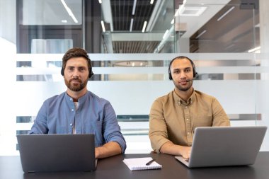 Kulaklık takan iki profesyonel ofisteki bir masada dizüstü bilgisayarla çalışıyor. Bu ayar, müşteri hizmetleri veya teknik destek, işbirliği ve profesyonel çalışma ortamını yansıtmaktadır..