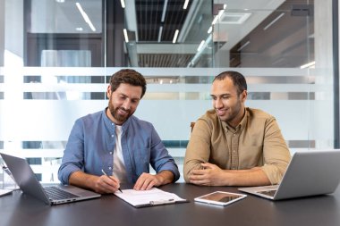 İki profesyonel bir ofiste iş birliği yapıyor. Biri kağıtları imzalarken diğeri onları gözden geçiriyor. Atmosfer, takım çalışması, işbirliği ve resmi iş etkileşimi öneriyor..