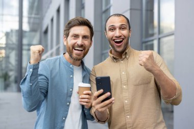 İki neşeli birey şehirsel bir alanda birlikte kutlama yaparken akıllı telefondan heyecan verici haberler izliyorlar. Bir kahve fincanını paylaşırken,.