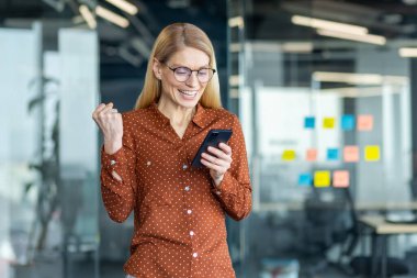 Profesyonel bir kadın ofis ortamında akıllı telefonundan olumlu haberler izlerken sevinir. Çevrede cam paneller ve renkli yapışkan notlar yer alıyor