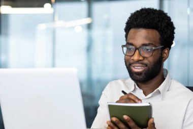 Ofiste oturup dizüstü bilgisayar üzerinde çalışırken not alan neşeli bir profesyonel. Kurulum üretkenlik, profesyonellik ve görevlerin yerine getirilmesine odaklanıyor