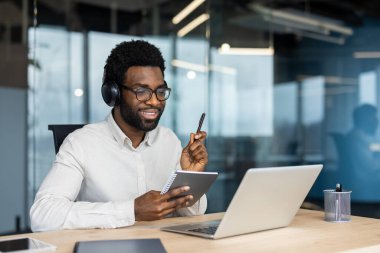 Modern bir iş yerinde dizüstü bilgisayarla video görüşmesi yapan, kulaklık takan, not defteri tutan, üretken iletişimi ve profesyonel odaklanmayı gösteren kendine güvenen bir birey.