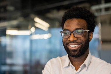 Gözlük takan neşeli genç bir adam, çağdaş bir ofis ortamında iken samimi bir şekilde gülümsüyor. Ruh hali pozitifliği, güveni ve profesyonelliği yansıtıyor..