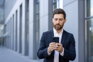 Profesyonel işadamı, kentsel bir caddede çağdaş bir ofis binasının önünde akıllı telefonu araştırıyor..