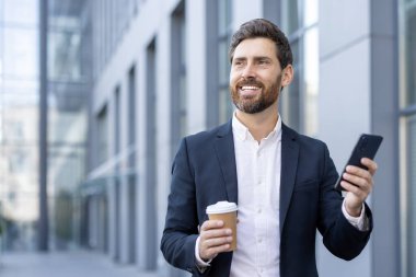 Elinde kahve fincanı ve akıllı telefon tutarken gülümseyen profesyonel bir adam. Kentsel arka plan modern, pozitif bir hava katıyor, başarıyı ve iletişimi simgeliyor.