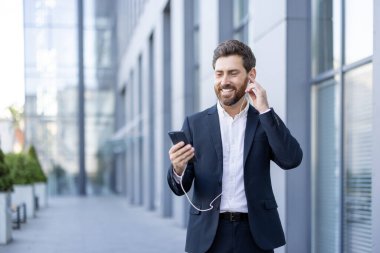 Takım elbise giyen kendine güvenen bir adam açık havada durur, akıllı telefon ve kulaklık kullanırken gülümser parlak bir şehir ortamında. Görüntü profesyonelliği, teknolojiyi ve olumlu bir çalışmayı temsil ediyor.