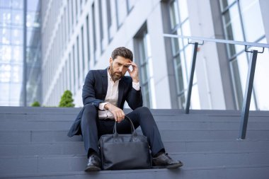 Resmi giyinmiş profesyonel bir adam modern bir binanın önünde düşünceli bir şekilde oturur ve tefekkür eder..