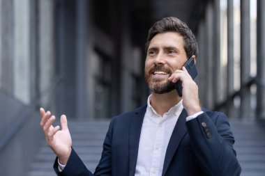 Çağdaş bir şehir ortamında akıllı bir telefonla konuşurken takım elbise giymiş, gülümseyen profesyonel bir birey. Görüntü başarıyı, iletişimi ve olumlu bir tutumu temsil ediyor..
