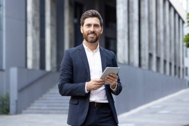 Kendine güvenen iş adamı resmi bir takım elbise giyiyor, açık havada tablet kullanıyor ve arka planı modern. Görüntü profesyonelliği, teknoloji kullanımını ve kurumsal bir ortamda hoş bir atmosferi gösteriyor.