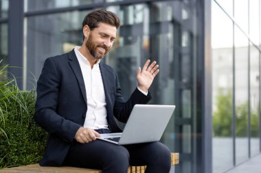 Takım elbiseli profesyonel bir adam dışarıda bankta otururken dijital olarak iletişim kuruyor, gülümsüyor..