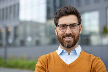 Şehir ortamında profesyonel bir şekilde gülümseyen, kendine güvenen gözlüklü bir adam. Arka planda modern mimari tarzda kentsel bir ortam sergileniyor