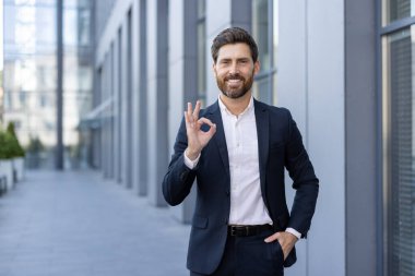 Takım elbise giyen profesyonel bir adam açık havada başarıyı, pozitifliği ve kendine güveni simgeleyen iyi bir el hareketi yapar. Modern kentsel arka plan, kurumsal ve kendine güvenen bir atmosferi geliştirir..