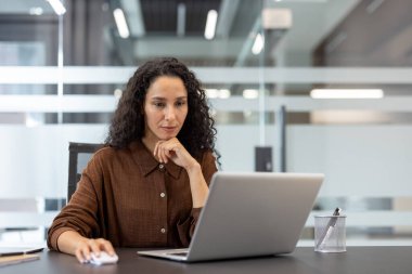 Cam kaplı bir ofiste dizüstü bilgisayar üzerinde çalışan, üretkenliği, odağı ve modern çalışma alanını temsil eden profesyonel bir kadın..