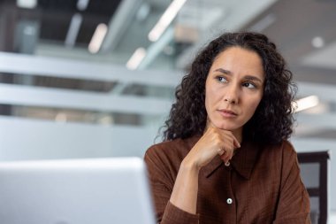 Kahverengi kıyafetli bir kadın, temiz ve modern bir çevreyle çevrili çağdaş bir ofiste masa başında özenle oturuyor. Evrak işleri ve problem çözmeyi sembolize eden bir dizüstü bilgisayarla..