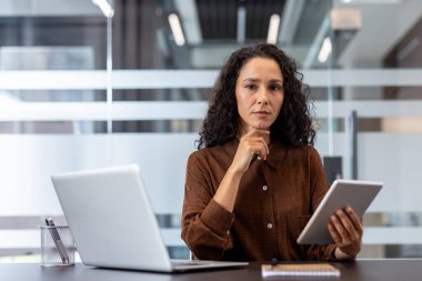 Profesyonel ofis ortamında kendine güvenen bir kadın düşünürken tablet ve dizüstü bilgisayar kullanıyor. Atmosfer, işbirlikçi bir çalışma alanında profesyonellik, modernlik ve odaklanma görevleri sunar..