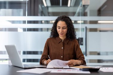 Kendine güvenen iş kadını, işyerindeki belgeleri özenle analiz ediyor, odaklanma ve profesyonellik gösteriyor. Modern ofis dekorasyonu, şirket atmosferi ve işe kendini adama.