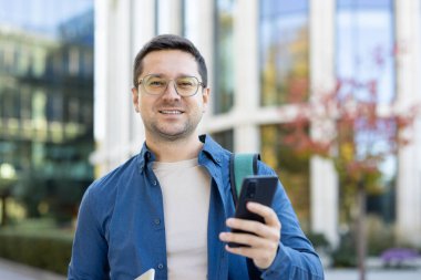 Dışarıda akıllı telefonlu neşeli genç bir adam, günlük kıyafetler giyiyor ve gülümsüyor..