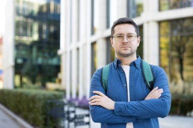 Gözlüklü ve sırt çantalı genç bir adam kampüs binasının önünde güven içinde poz veriyor. Kurulum modern, kentsel bir eğitim atmosferini yakalıyor