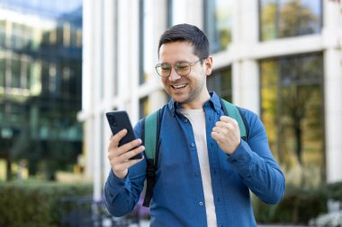 Gözlüklü ve sırt çantalı neşeli genç bir adam telefonuna bakarken gülümsüyor, arka planda modern mimari olan bir kampüs ortamında eğlencesini ifade ediyor..