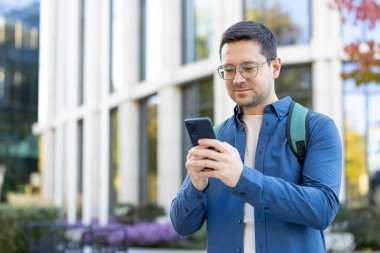 Gözlüklü ve sırt çantalı bir adam modern mimarinin yanında akıllı telefonunu kullanıyor..