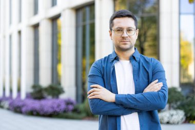 Açık havada kollarını kavuşturmuş kendinden emin bir gencin portresi. Modern mimari ve peyzaj çevresi, profesyonelliği yansıtan çağdaş bir atmosfer sağlıyor.