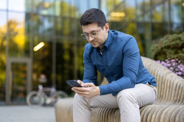 Genç bir adam açık havada ahşap bir bankta oturuyor ve telefonuna odaklanmış bir şekilde bakıyor. Etrafında modern mimari, yeşillik ve rahat bir ortam ile manzaralı bir şehir ortamı var..