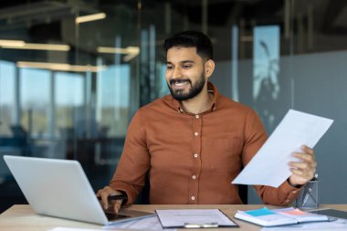 Çağdaş bir ofis ortamında dizüstü bilgisayar üzerinde çalışırken belgelere bakıp, üretkenlik ve profesyonelliği aktaran gülümseyen iş adamları.