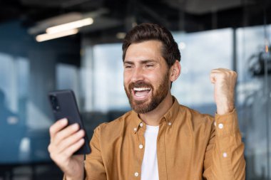 Neşeli bir adam heyecan ve mutluluk sergiliyor. Akıllı telefonunu modern ofis ortamında muzaffer bir şekilde tutuyor..