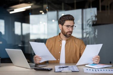 Gözlüklü ve sıradan gömlekli profesyonel bir adam ofis ortamında belgeleri inceliyor. Masanın üzerinde bir dizüstü bilgisayar ve çizelgeler var. Çevre, modern ve dinamik bir iş yeri öneriyor.